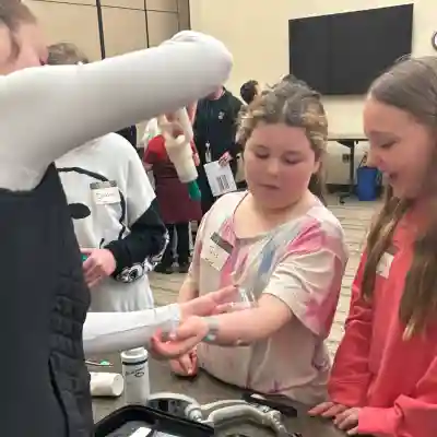 Two students lean in attentively as a healthcare professional demonstrates a medical model. Various medical supplies and educational materials are displayed on the table