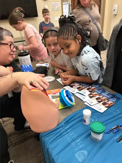 Two students lean in attentively as a healthcare professional demonstrates a medical model. Various medical supplies and educational materials are displayed on the table