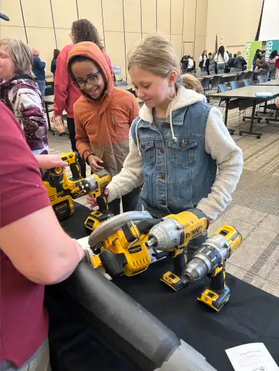 Two students interact with power tools on a display table, guided by a volunteer, while other students and attendees engage in activities in the background