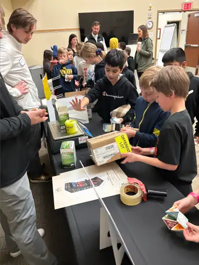 [Image description: A group of students gathers around a table with various packaging materials and equipment, interacting with volunteers and exploring logistics tools.